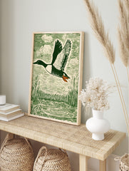 Green woodcut-style print of a flying duck with spread wings over water and reeds, in an alternate framed mockup