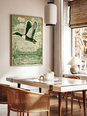 Green woodcut-style print of a flying duck with spread wings over water and reeds, in a styled room mockup