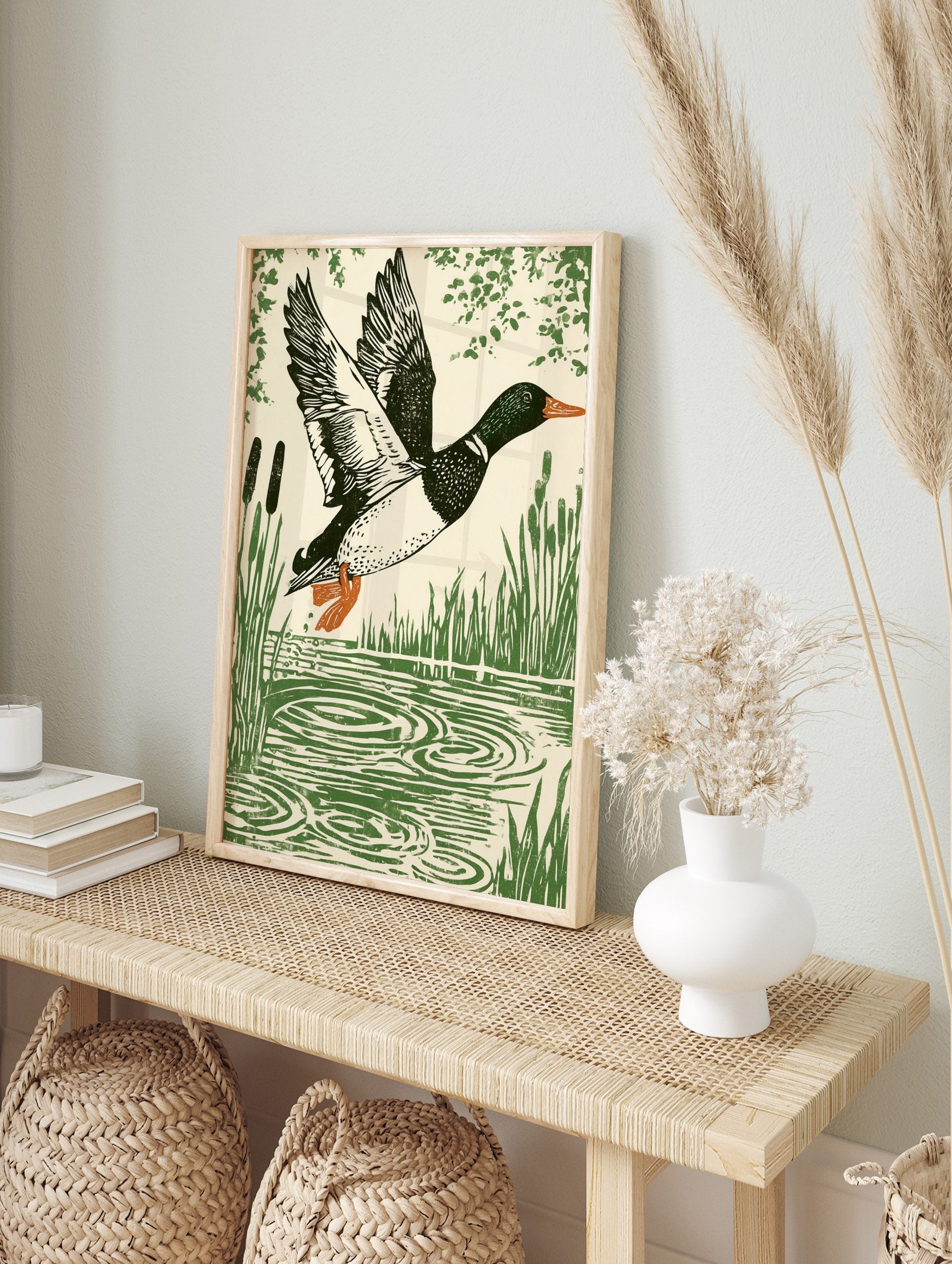 Linocut style print of a mallard duck in flight over water with reeds and foliage, in an alternate framed mockup
