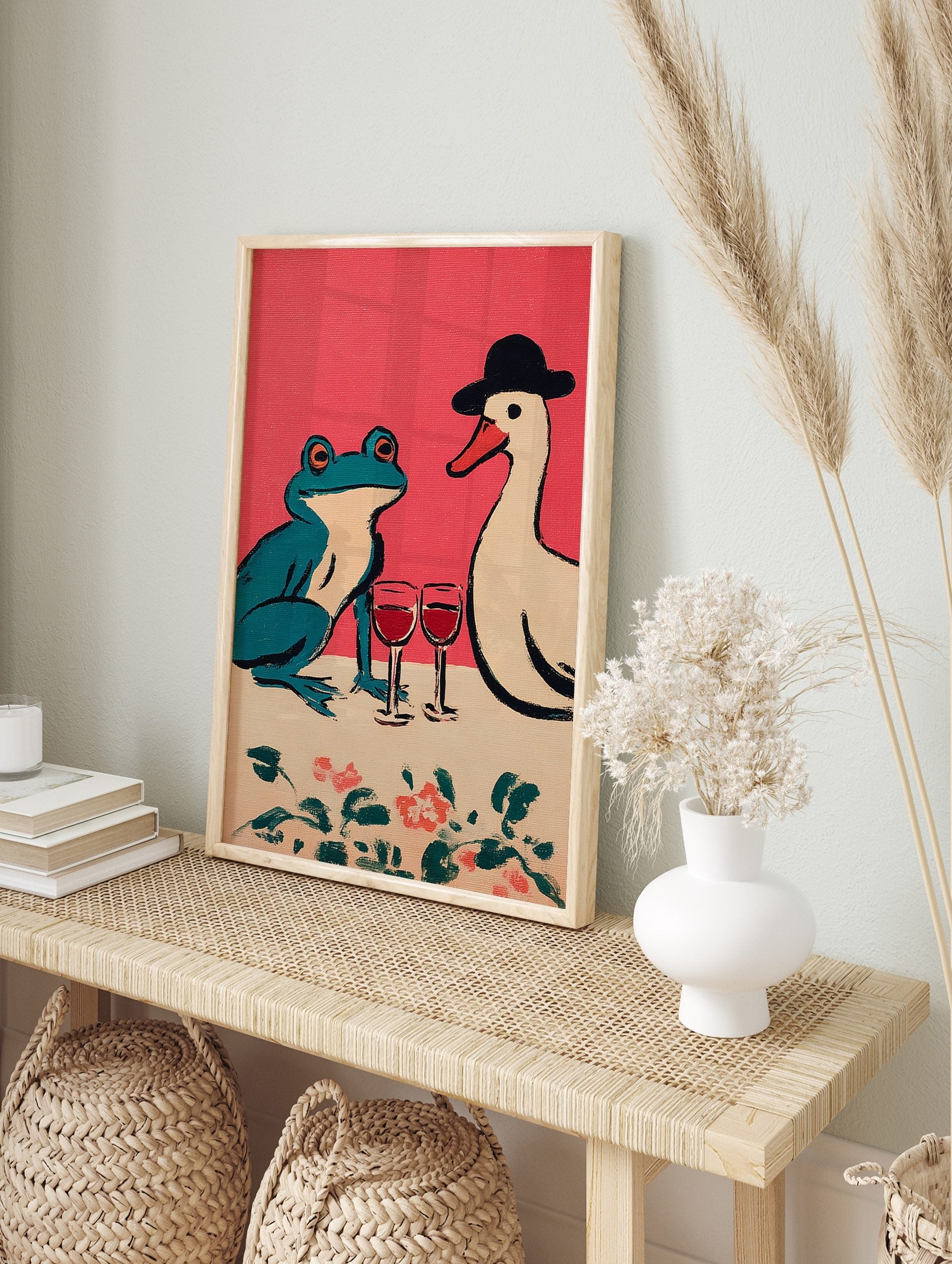 Illustrative print of a teal frog and a white duck in a hat sharing red wine on a pink, in an alternate framed mockup