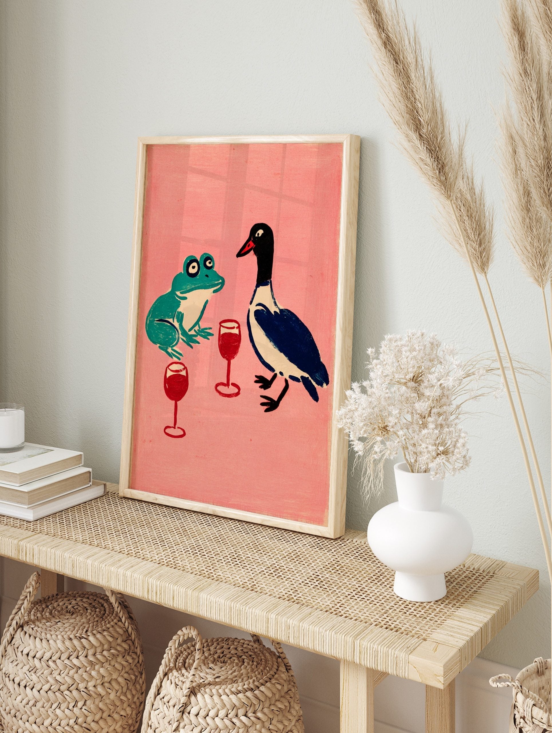 Illustrative print of a green frog and blue duck with red wine glasses on a pink background, in an alternate framed mockup