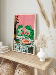 Whimsical painting of two green frogs on a dinner date with wine and food, against a pink, in an alternate framed mockup
