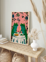 Two whimsical green frogs on a date at a table with wine and candle, pink background with, in an alternate framed mockup