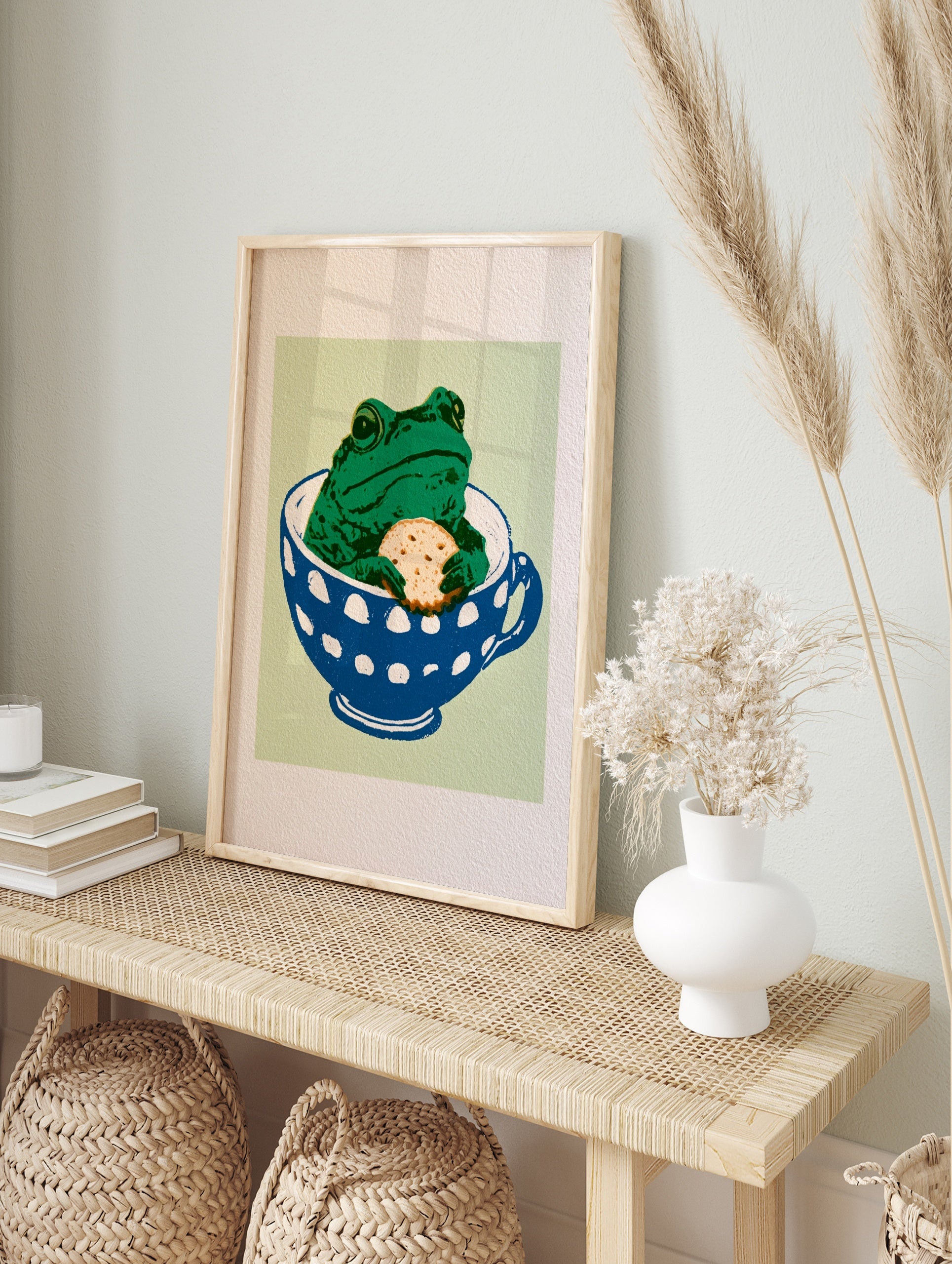 Whimsical green frog sitting in a blue and white polka-dot teacup holding a biscuit on a, in an alternate framed mockup
