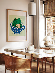 Whimsical green frog sitting in a blue and white polka-dot teacup holding a biscuit on a light, in a styled room mockup