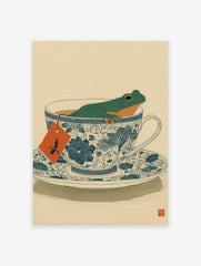 Green frog relaxing in a blue and white patterned teacup, with an orange tea bag tag, on a light beige background.