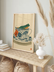 Green frog relaxing in a blue and white patterned teacup, with an orange tea bag tag, on a light beige background. - Image 3