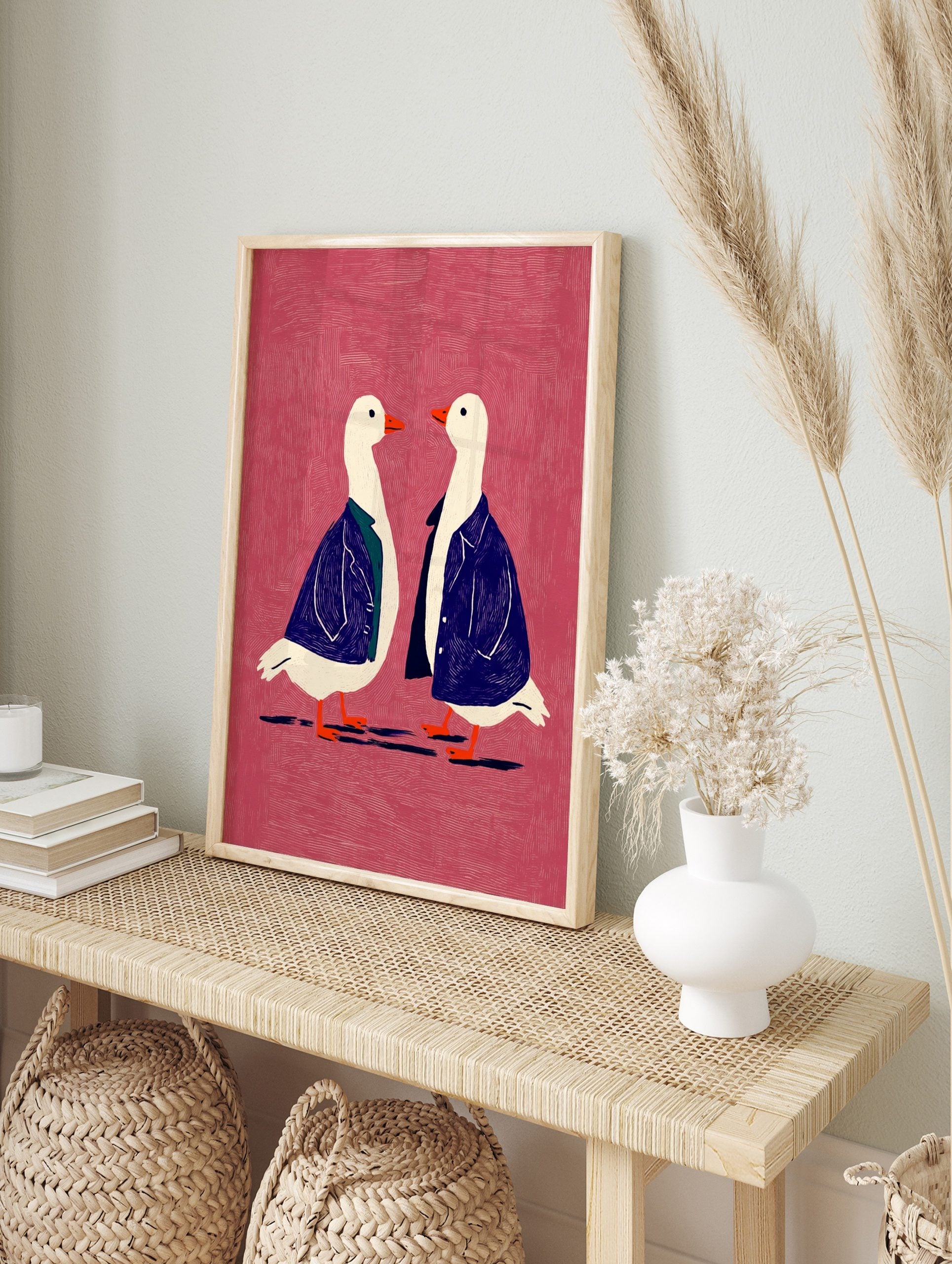 Two white geese in blue jackets with orange beaks on a textured pink background, facing each, in an alternate framed mockup