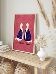 Two white geese in blue jackets with orange beaks on a textured pink background, facing each, in an alternate framed mockup