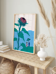Illustrative print of a giant teal and pink thistle flower towering over two small figures, in an alternate framed mockup