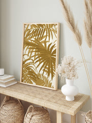 Gold palm leaf tropical botanical wall art print with overlapping foliage on a light background, displayed as the same artwork in an alternate framed wall art mockup