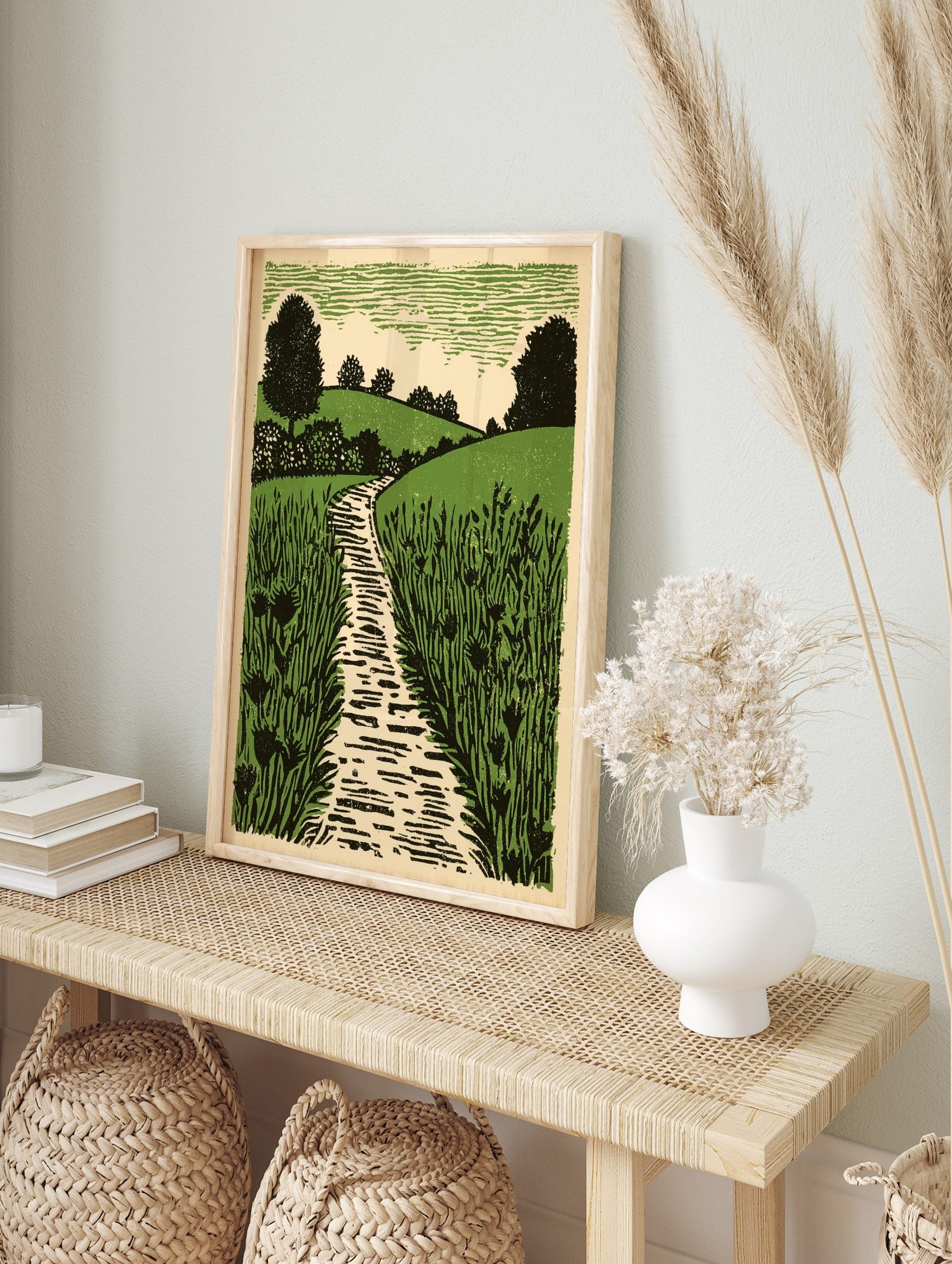 Linocut style print of a winding path through green fields, rolling hills, and trees under a, in an alternate framed mockup