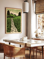 Linocut style print of a winding path through green fields, rolling hills, and trees under a, in a styled room mockup