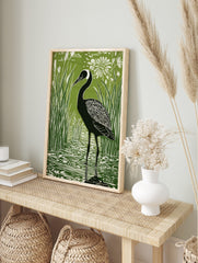 Green and white linocut style print of a black crane bird in reeds and water with a white, in an alternate framed mockup