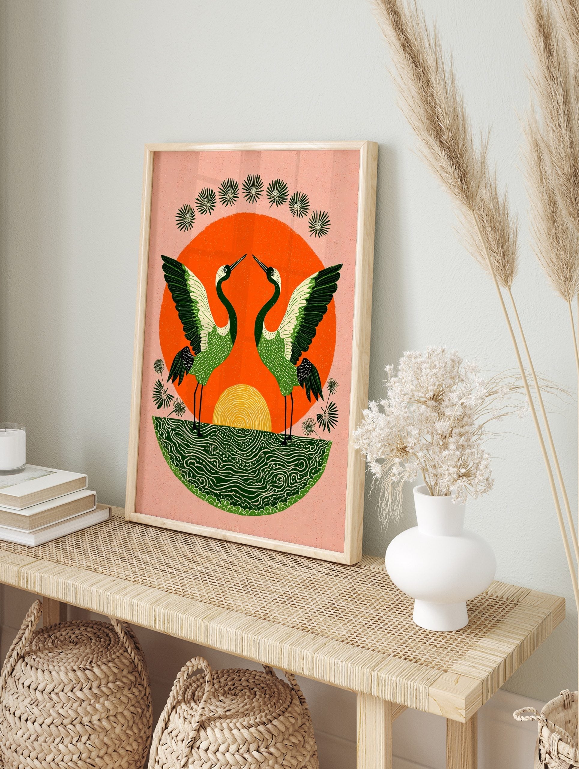 Illustrative print of two green cranes with spread wings against an orange sun and green water, in an alternate framed mockup