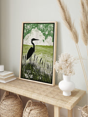 Green linocut style print of a heron in silhouette standing in tall reeds by water, with, in an alternate framed mockup