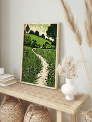 Green landscape woodcut style print with winding path, tall grass, small flowers, and, in an alternate framed mockup