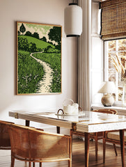Green landscape woodcut style print with winding path, tall grass, small flowers, and tree-covered, in a styled room mockup