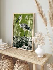 Green linocut print of a patterned stork wading in water with reeds and reflections, in an alternate framed mockup