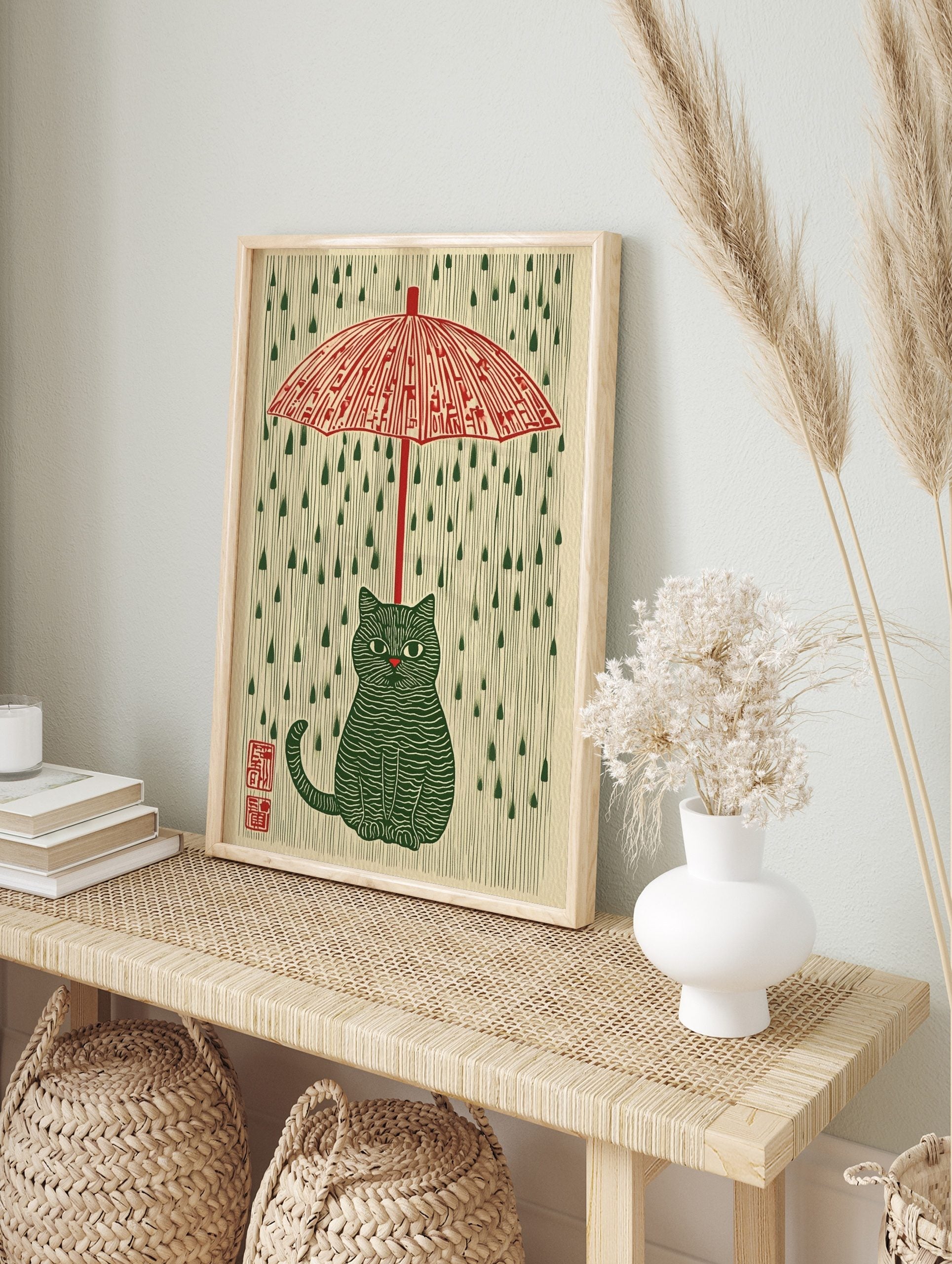Illustrative green striped cat under a red umbrella with green raindrops on a cream background, in an alternate framed mockup