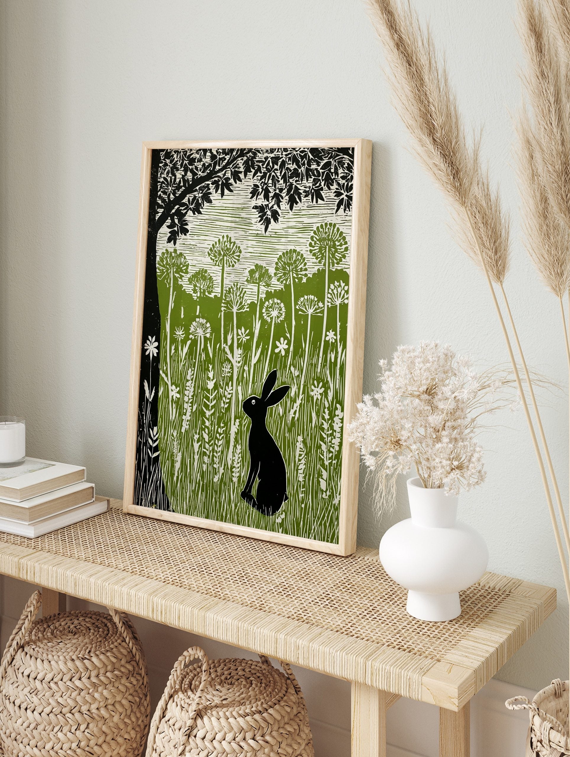 Linocut print of a black hare in a green meadow with tall white wildflowers and a large black, in an alternate framed mockup