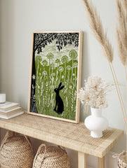 Linocut print of a black hare in a green meadow with tall white wildflowers and a large black, in an alternate framed mockup