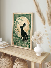 Black hare silhouette against a gold speckled moon, surrounded by green foliage in a linocut, in an alternate framed mockup