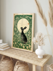Woodcut print of a black hare looking up at a large cream moon with green sky and white, in an alternate framed mockup
