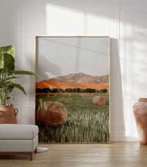 Photographic print of a field with hay bales, green grass, a row of trees, orange hills, and mountains under a grey sky. - Image 3