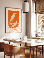 White line art illustration of a heron standing on large plants against a vibrant orange background, in a styled room mockup