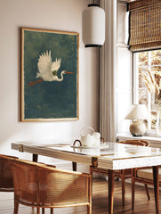 Illustrative print of a white heron flying horizontally with a green frog on its back against a, in a styled room mockup