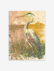 Illustrative art print of a heron bird standing in tall grasses by water, with a muted pink and gold sky