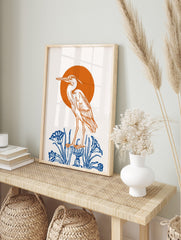 Illustrative heron bird standing on blue pedestal with blue plants, orange sun background, in an alternate framed mockup