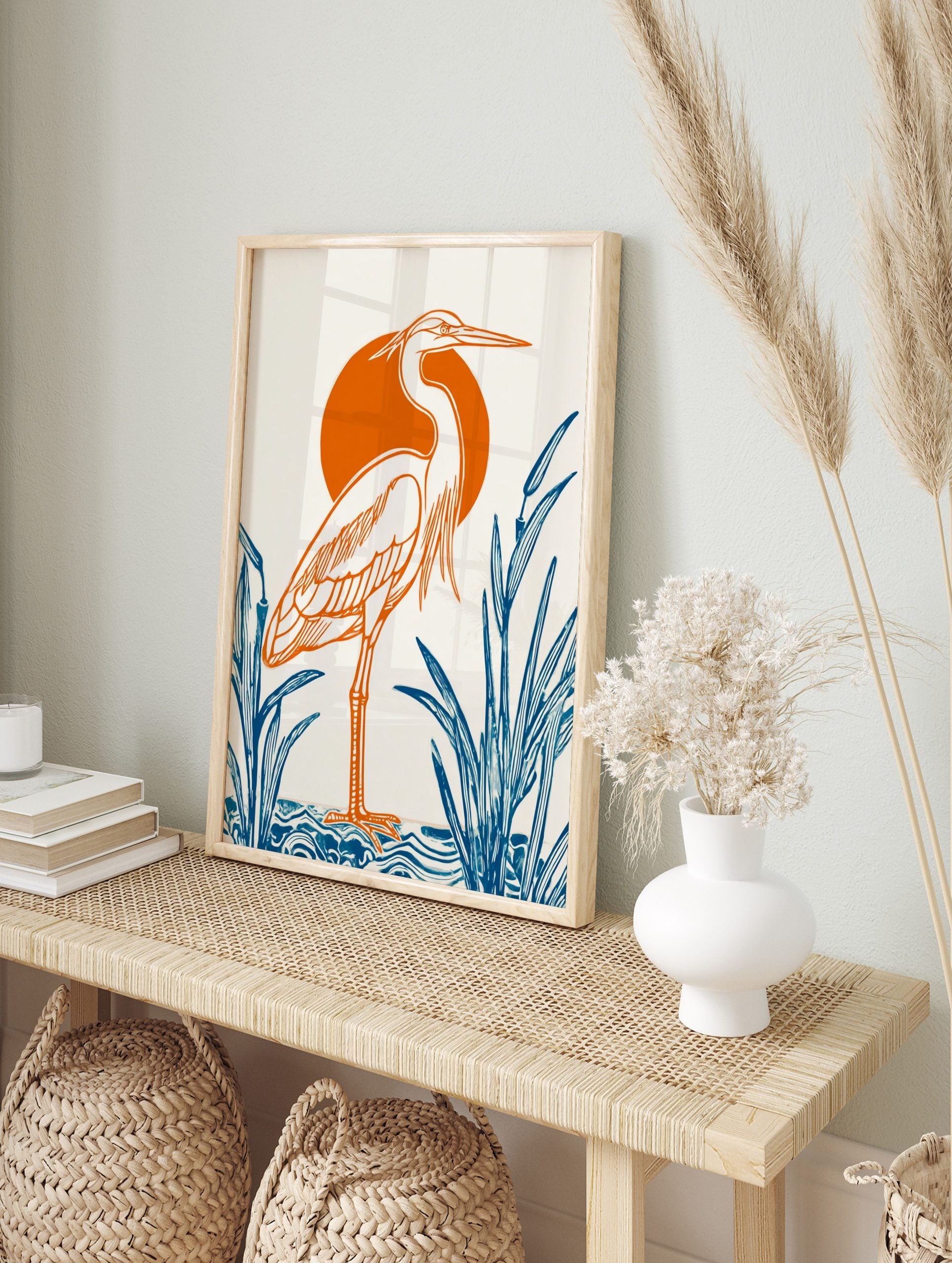 Illustrative heron bird print with orange outline, orange sun, and blue reeds on white, in an alternate framed mockup