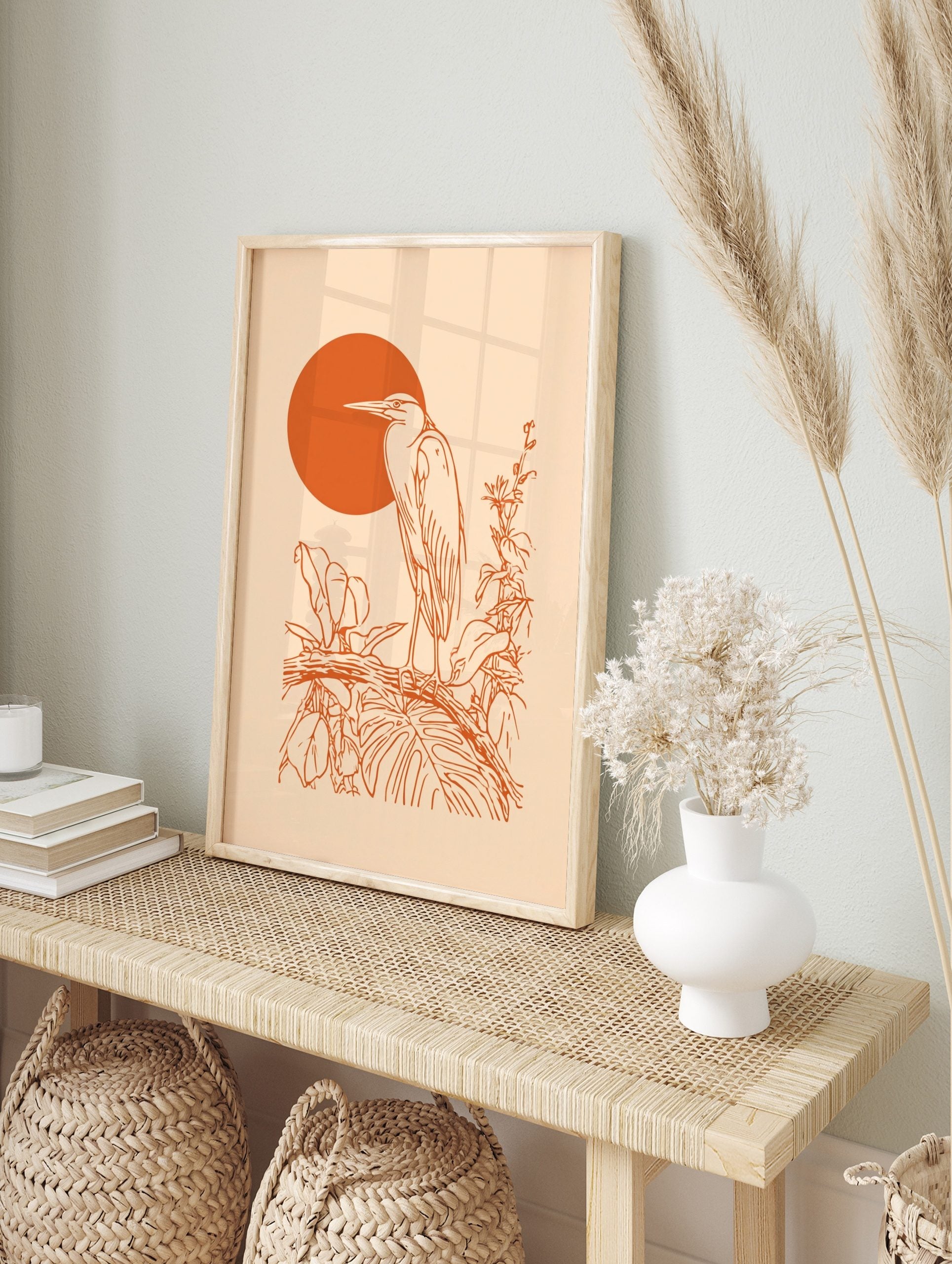 Orange line art of a heron bird on a branch with tropical leaves and a large orange sun on a, in an alternate framed mockup