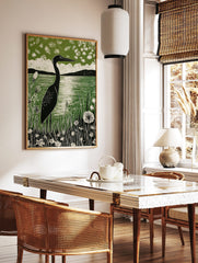 Linocut print of a black heron by a green lake, surrounded by white wildflowers and distant hills, in a styled room mockup
