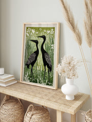 Linocut print of two black herons standing in a vibrant green wetland with white reeds and, in an alternate framed mockup