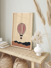 Illustrative red and cream hot air balloon floating above abstract brown and dark grey hills, in an alternate framed mockup
