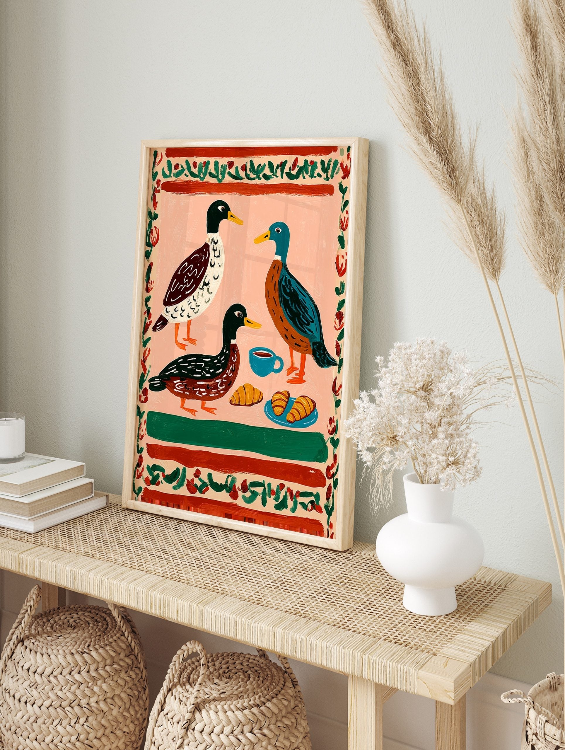 Illustrative print of three ducks, coffee cup, and croissants on a pink background with a red, in an alternate framed mockup