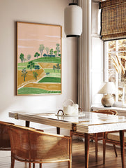 Illustrative landscape art print with green hills, gold fields, scattered trees, and a peach sky, in a styled room mockup