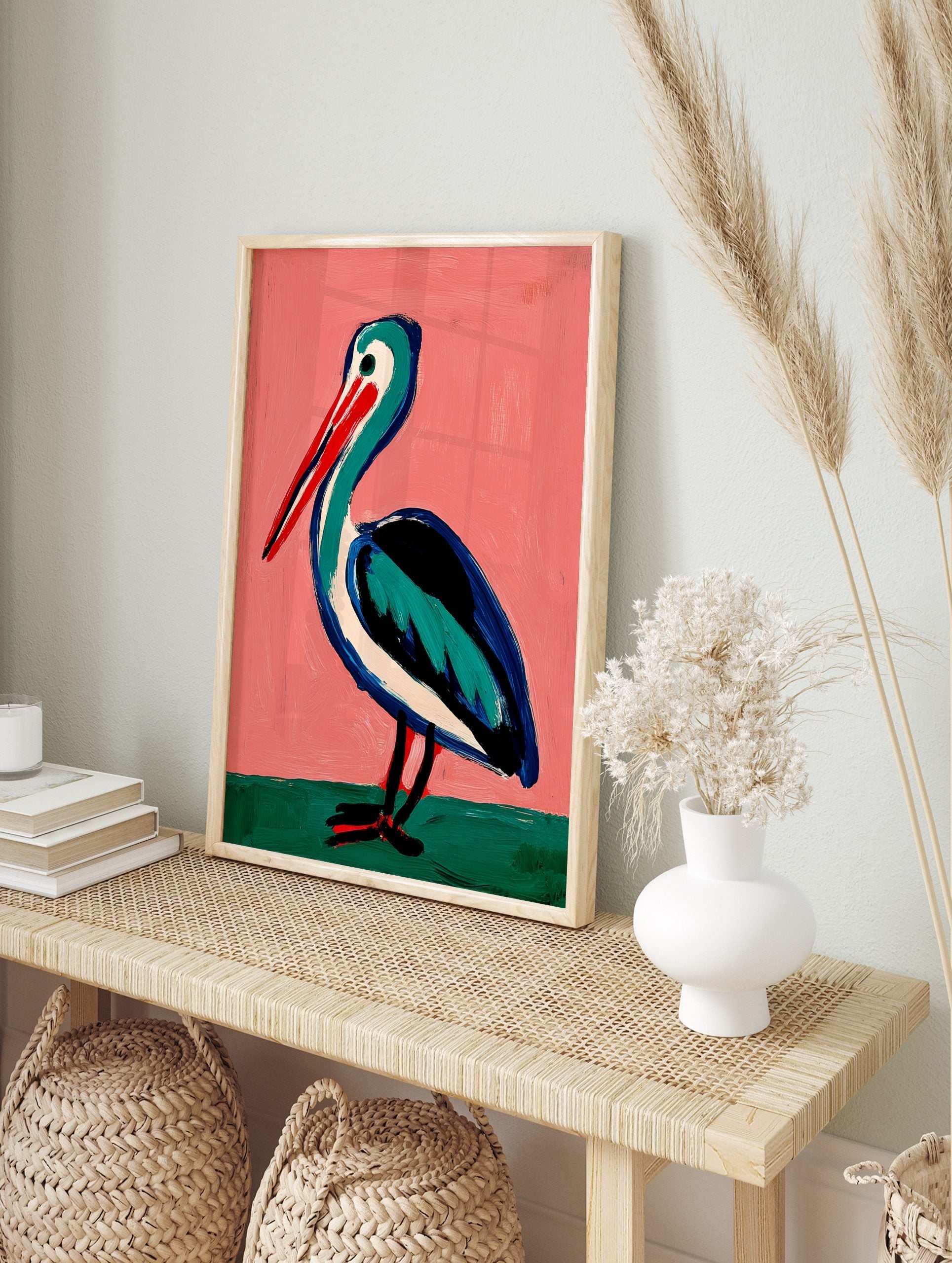 Illustrative painting of a pelican with a red beak, teal and blue body on a pink background, in an alternate framed mockup