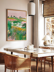 Illustrative landscape art print with green hills, pink sky, a red-roof house, and foreground, in a styled room mockup