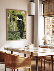 Linocut-style print of a black and white kingfisher bird on a branch over green water with reeds, in a styled room mockup
