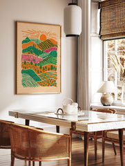 Illustrative layered landscape art print with orange sun, green hills, magenta fields, and trees, in a styled room mockup