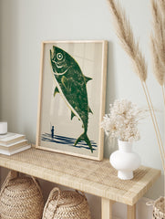 Illustrative print of a large green fish leaping from blue water, with a small fisherman, in an alternate framed mockup