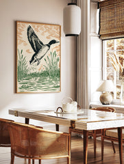 Linocut style art print of a mallard duck flying from water with green reeds and orange-brown sky, in a styled room mockup