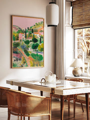 Illustrative Mediterranean landscape print with pink houses, green hills, cypress trees, and a, in a styled room mockup