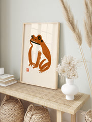 Minimalist frog illustration in brown brush strokes with a red seal on a cream background art, in an alternate framed mockup