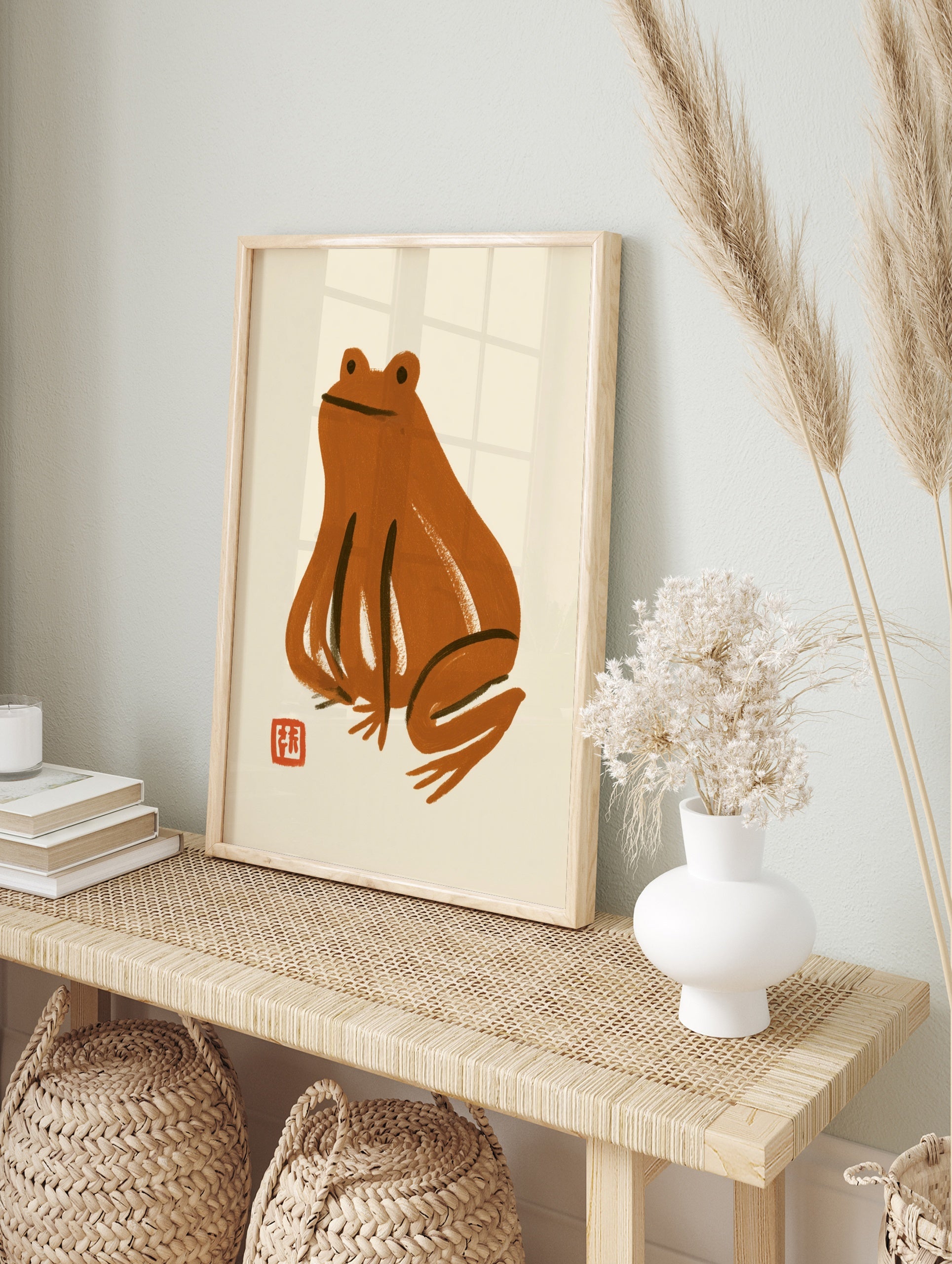 Minimalist brown frog illustration with a small red stamp on a cream background, illustrative, in an alternate framed mockup