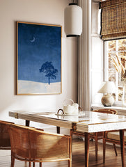 Minimalist wall art print of a single tree and crescent moon over a snowy landscape in deep blue, in a styled room mockup